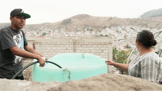 Zwei Menschen bei einem Wasserkübel aus Plastik mit Blick auf eine Stadt. © Screenshot 