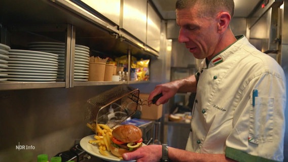 Ein Mann schüttet Pommes frittes auf einen Teller, auf dem schon ein Burger liegt. © Screenshot 