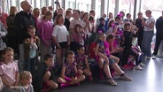 Bunt und pink angezogene Kinder haben sich für ein Gruppenfoto aufgestellt. © Screenshot 