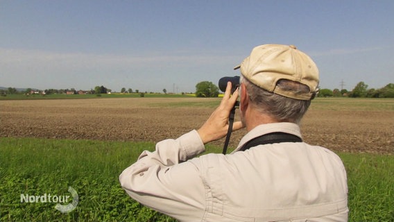 Ein Mann in beigem Hemd steht auf einem Feld und schaut durch ein Fernglas. © Screenshot 