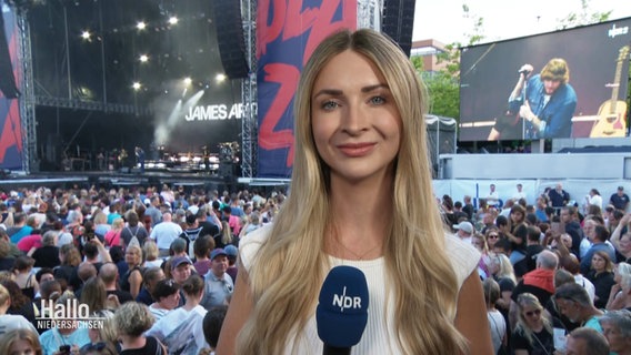 Reporterin Kerstin Liegendza, hinter ihr ist eine große Menschenmenge vor einer Bühne zu sehen. © Screenshot 