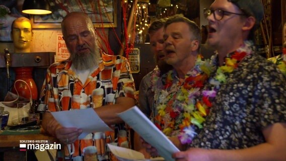 Männer in Hawaii-Hemden und künstlichen Blumenketten stehen dicht beieinander und singen. In ihren Händen halten sie Papier, auf dem offenbar der Liedtext steht. © Screenshot 