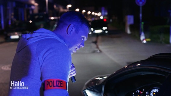 Ein Polizist in Zivil mit einer roten Armbinde leuchtet mit einer Tachenlampe in ein Auto. © Screenshot 