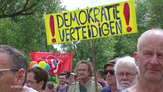 "Demokratie verteidigen" steht auf einem gelben Schild, das während einer Demonstration über die Köpfe der Teilnehmer ragt. © Screenshot 