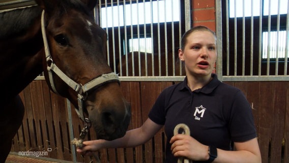 Eine junge Frau steht in einem Pferdestall und hält ein Pferd am Zügel fest. © Screenshot 