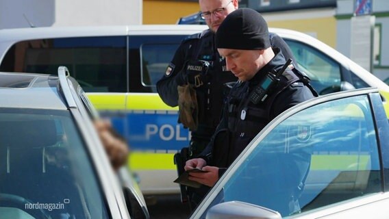 Ein Polizeibeamter steht vor einer geöffneten Autotür und kontrolliert die Papiere des Fahrers. © Screenshot 