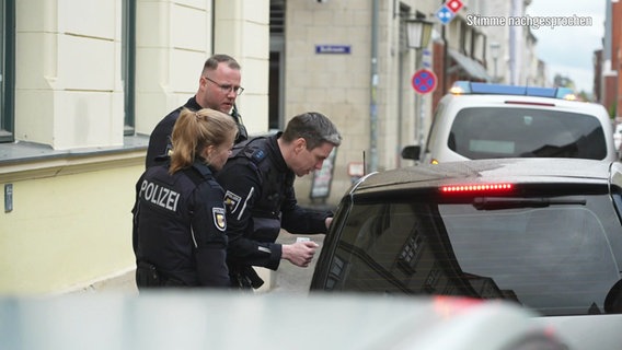 Drei Polizisten stehen neben einem Kleinwagen in einer Straße in Stralsund. Ein Polizist beugt sich zum Fenster auf der Fahrerseite hinunter. © Screenshot 