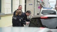 Drei Polizisten stehen neben einem Kleinwagen in einer Straße in Stralsund. Ein Polizist beugt sich zum Fenster auf der Fahrerseite hinunter. © Screenshot 