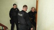 Drei Polizisten stehen in Stralsund in einem Treppenhaus vor einer verschlossenen Wohnungstür. © Screenshot 