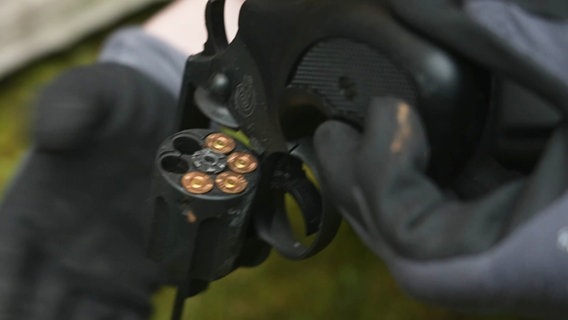 Zwei Hände mit dunkel Handschuhen halten eine Pistole, die Trommel der Schreckschusswaffe ist herausgeklappt, es sind vier Patornen zu sehen. © Screenshot 