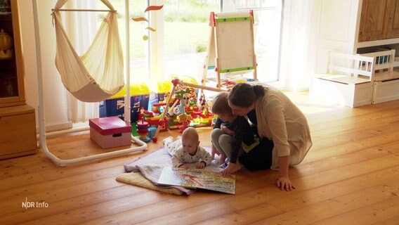 Eine Mutter sitzt mit einem Kleinkind und einem Baby auf dem Boden und schaut gemeinsam mit den Kindern eine bunte Karte an. Hinter ihnen stehen vcerschiedene Spielsachen. © Screenshot 