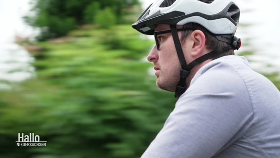 Ein Mann fährt Fahrrad. Er trägt einen weißen Helm und graue Kleidung, hinter ihm zieht verschwommen die Landschaft vorbei. © Screenshot 