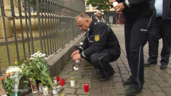 Blumensträuße und Kerzen liegen auf dem Boden vor einem Zaun. Zwei Polizeibeamte hocken bzw. stehen davor und entzünden weitere Kerzen. © Screenshot 
