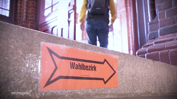 Auf einer Treppenstufe klebt ein roter Zettel mit einem wegweisenden Pfeil und der Aufschrift "Wahlbezirk". © Screenshot 