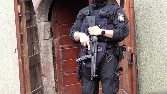 Ein Polizeibeamter mit Maschinengewehr vor einem Hauseingang. © Screenshot 