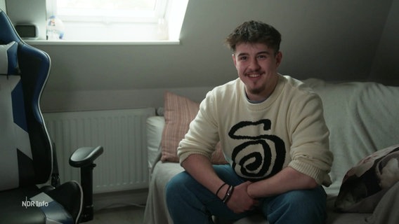 Der 17-Jährige Angelos Andreadis sitzt in seinem Zimmer auf einem Sofa und lächelt in die Kamera. Er hat kurze Braune Haare, trägt Jeans und einen hellen Wollpulli. © Screenshot 