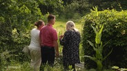 Drei Personen stehen an einer Hecke in einem sehr grünen Garten. © Screenshot 