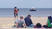 Mehrere Menschen sitzen am Strand und schauen auf das Meer. © Screenshot 