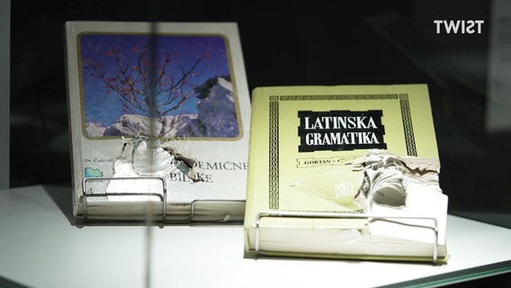 In einer Glasvitrine liegen zerschossene Schulbücher. © Screenshot 