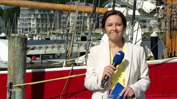 Romy Hiller moderiert draußen am Museumshafen in Flensburg. Hinter ihr liegt ein altes Segelschiff im Wasser. © Screenshot 