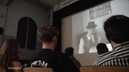 Jugendliche sitzten vor einer Kinoleinwand, auf die ein Film projeziert wird. Der Film ist in schwarz/weiß und es ist ein Jugendlicher zu sehen, der wie ein Film Noir Detektiv gekleidet ist. © Screenshot 
