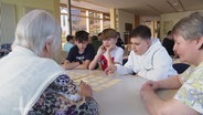 Jugendliche und Seniorinnen sitzen an einem Tisch und spielen gemeinsam. © Screenshot 