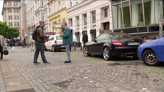 Zwei Männer unterhalten sich auf einem Platz. © Screenshot 