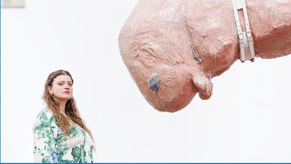 Sophie Süßmilch steht neben einem ihrer Kunstwerke: Eine organisch wirkende, fleischfarbene Skulptur eines Wesens, das an Stahlringen von der Decke hängt. © Screenshot 