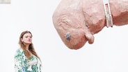 Sophie Süßmilch steht neben einem ihrer Kunstwerke: Eine organisch wirkende, fleischfarbene Skulptur eines Wesens, das an Stahlringen von der Decke hängt. © Screenshot 