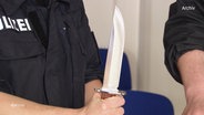 Ein Polizeibeamter hält ein großes Messer. (Archivfoto) © Screenshot 