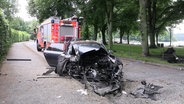 Ein stark zerstörtes Auto steht am Straßenrand, im Hintergrund ein Einsatzwagen der Feuerwehr. © Screenshot 
