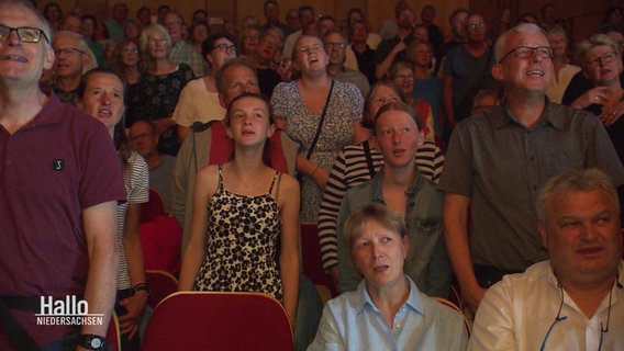 Das Publikum eines Konzerthauses erhebt sich zum gemeinsamen Singen von den Plätzen. © Screenshot 