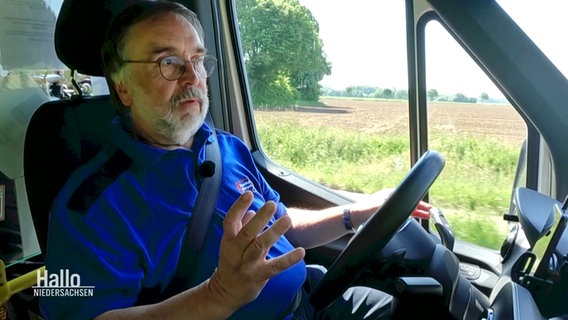 Ein Mann mit grauem Bart und blauem Shirt sitzt am Steuer eines Klein-Busses. © Screenshot 