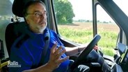 Ein Mann mit grauem Bart und blauem Shirt sitzt am Steuer eines Klein-Busses. © Screenshot 