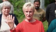 Ein Gruppe von Menschen steht hinter und neben einer Frau, die fragend die Hand hebt. © Screenshot 
