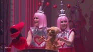 Zwei als Außerirdische verkleidete Frauen mit Puppen auf einer Bühne. © Screenshot 