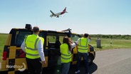 Drei Personen mit Warnwesten beobachten ein Flugzeug bei der Landung. © Screenshot 
