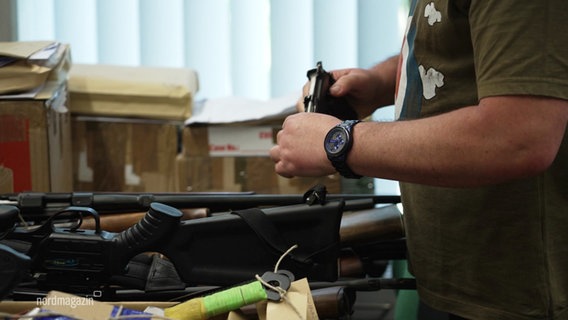 Waffen auf einem Tisch und eine Person mit einer davon in der Hand. © Screenshot 