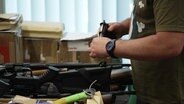 Waffen auf einem Tisch und eine Person mit einer davon in der Hand. © Screenshot 