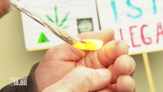 Nahaufnahme: Eine Person zündet einen Joint an. Im Hintergrund sind Protestplakate zum Thema Camnnabis-Legalisierung zu sehen. © Screenshot 