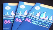 Werbesprospekte der Warnemünder Woche liegen auf einem Tisch. © Screenshot 