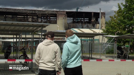 Zwei Personen vor einer ausgebrannten Lagerhalle. © Screenshot 
