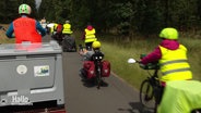 Eine Fahrradkolonne mit viel Gepäck fährt einen Weg zwischen Bäumen entlang. © Screenshot 