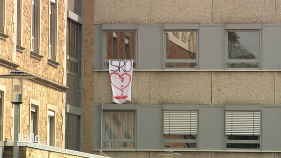 Aus einem Fenster hängt ein mit den Buchstaben SRO und einem Herzen besprühtes Laken. © Screenshot 
