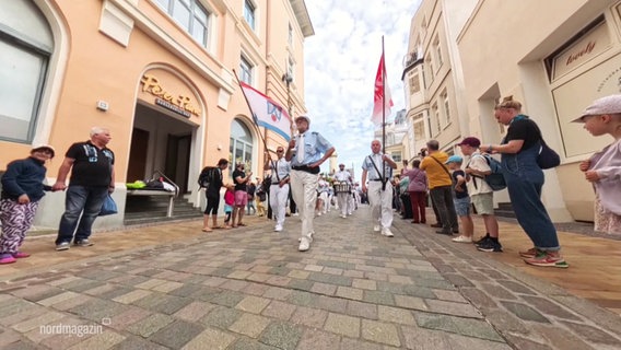 Uniformierte Männer mit Flaggen in den Händen führen eine Parade durch die Warnemünder Innenstadt an. © Screenshot 