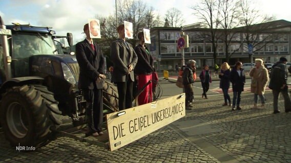 An einem Traktor sind drei als Politiker der Bundesregierung verkleidete Puppen befestigt. © Screenshot 