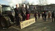 An einem Traktor sind drei als Politiker der Bundesregierung verkleidete Puppen befestigt. © Screenshot 
