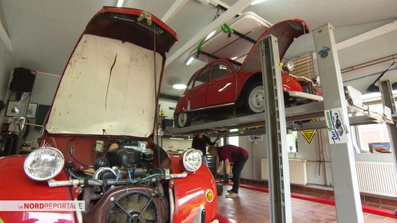 Ein Mann und eine Frau reparieren eine Ente auf einer Hebebühne in einer Autowerkstatt. © Screenshot 