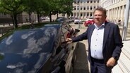 Gesundheitsminister Philipi steht neben seinem Diesel betriebenen Dienstwagen und erklärt dessen Vorzüge für sich. © Screenshot 
