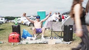Ein Mann mit Sonnenbrille sitzt auf einem Campingstuhl auf dem Campinggelände des Airbeat One © Screenshot 